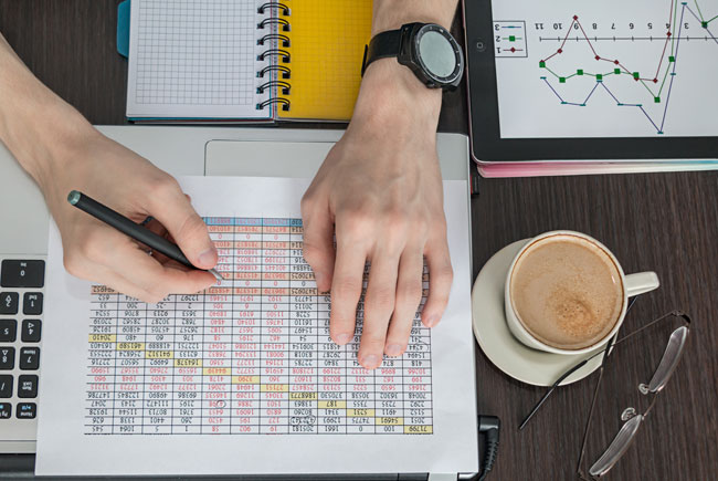 person working on a spreadsheet on a desk with a cup of coffee, glasses and an iPad with a graph